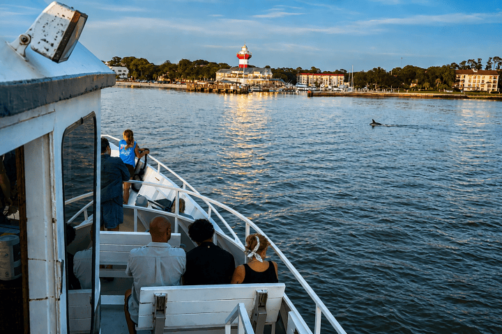 People on a boat near a lighthouse by the shore at sunset.