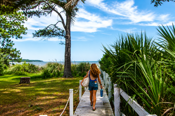 Woman walking on a wooden path towards the beach, surrounded by trees and greenery.