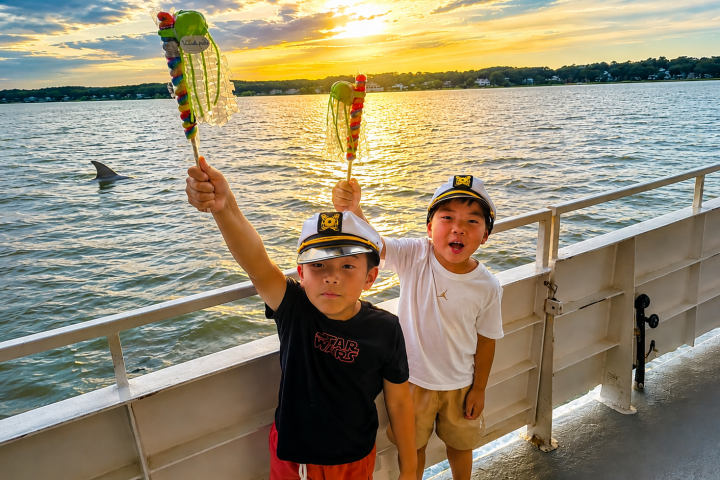 Two kids on a boat with toy swords at sunset, water and distant shoreline visible.