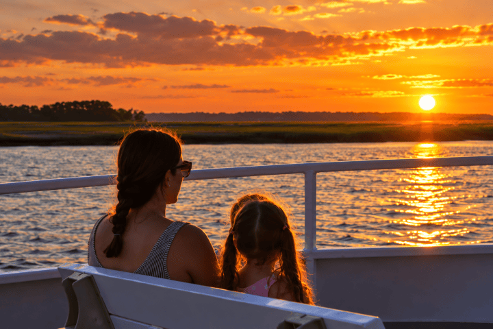 Woman and child on boat at sunset with orange sky and water reflections.