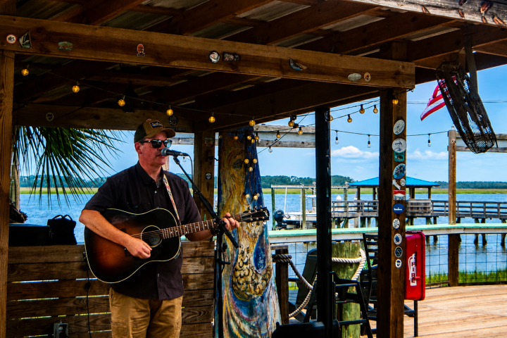 Man playing guitar and singing on a wooden deck by the water.
