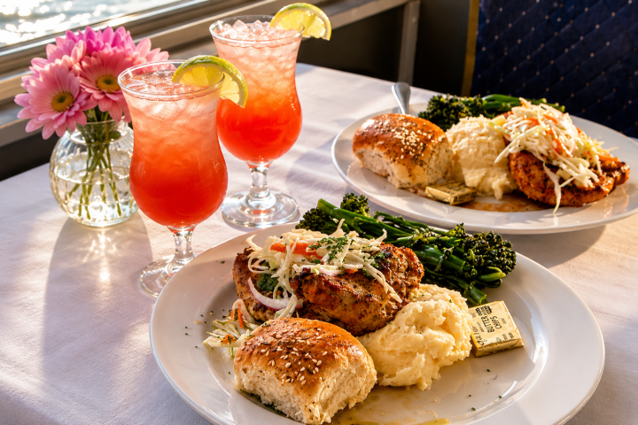 Two plates of meals with drinks by a window, featuring chicken, mashed potatoes, and buns.
