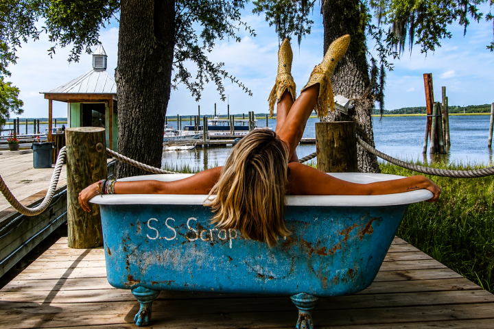 Person in a blue bathtub on a dock, wearing boots, facing a waterfront view.
