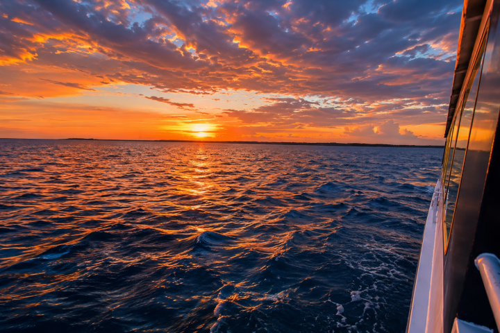 Boat on ocean at sunset with vibrant orange sky and clouds.