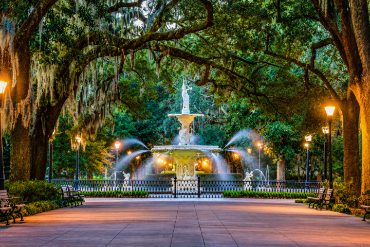 Lit fountain with water streams in a park framed by large trees and benches.