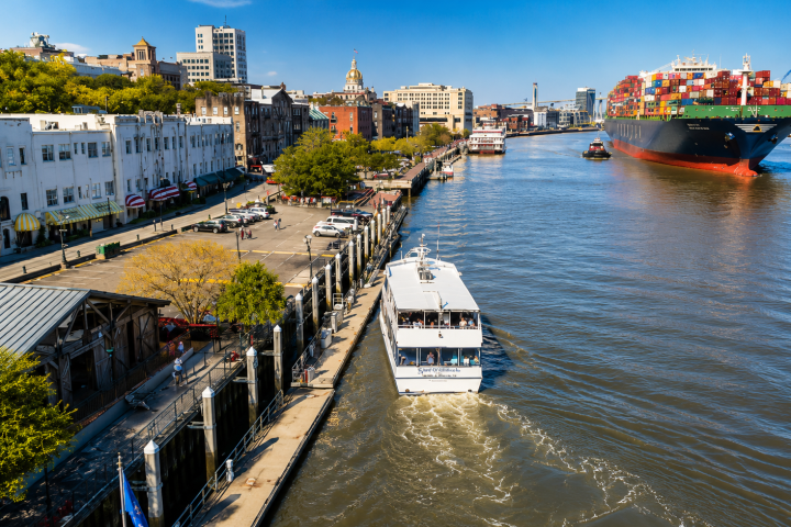 River scene with cargo ship and tour boat near city buildings.