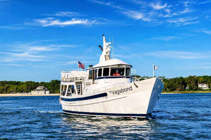 White ferry boat named 'Vagabond' sailing on clear blue water with trees in the background.