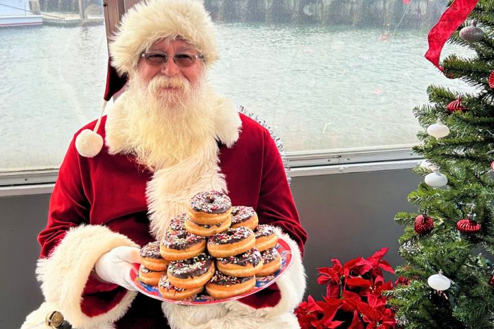 Santa Claus holds a plate of donuts by a decorated Christmas tree on a boat.