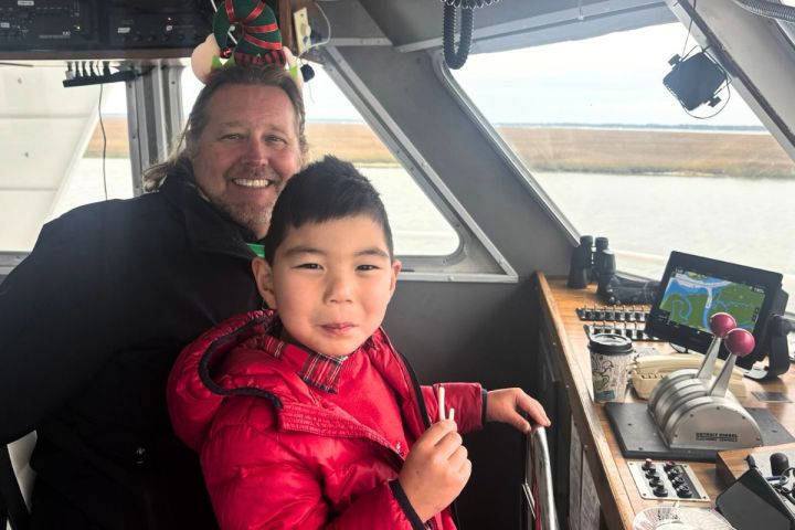 Smiling man and child in red coat on a boat's control deck with water view.
