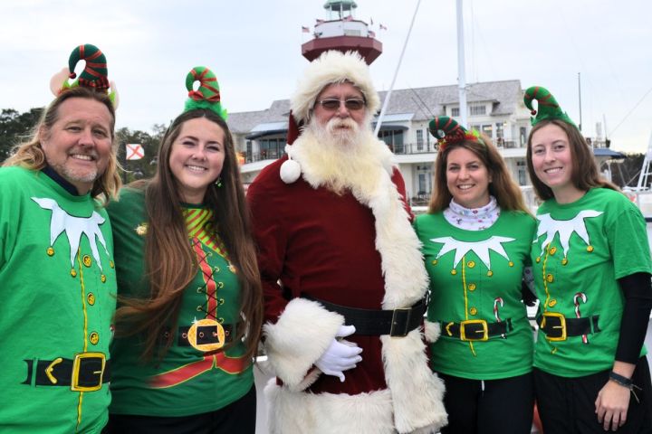 Santa with four people in elf costumes near a harbor with boats in the background.