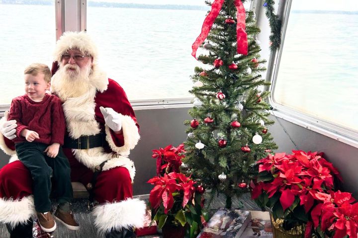 Santa with child on lap next to a Christmas tree and poinsettias on a boat.
