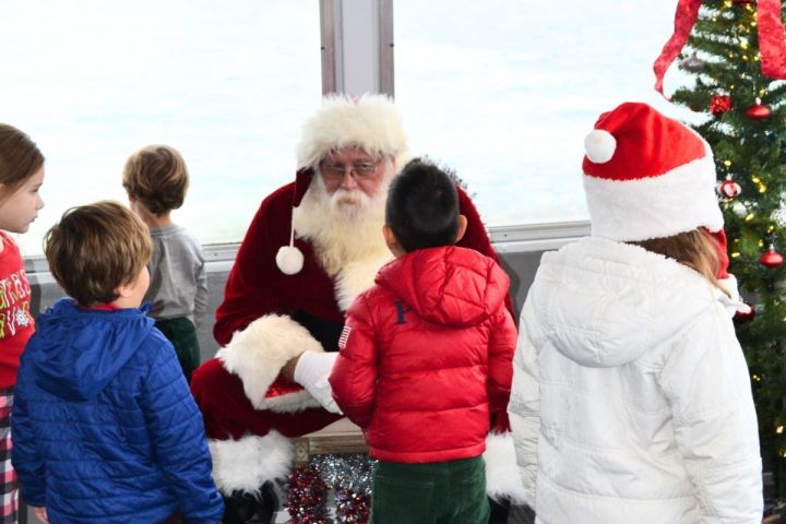Santa Claus sitting with children around a decorated Christmas tree indoors.