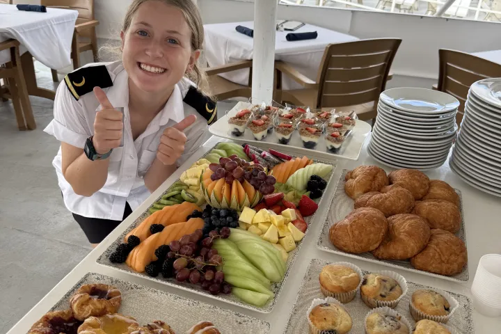 Smiling person in uniform giving thumbs up near trays of pastries and fruit on a boat.