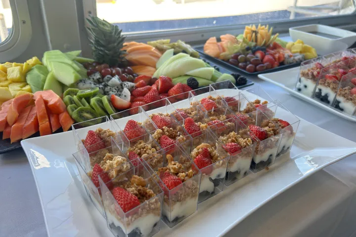 Trays with assorted fruits and yogurt parfaits topped with granola and strawberries on a table.