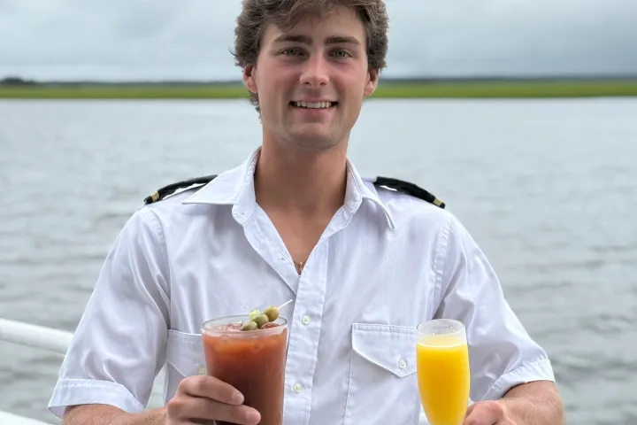 Man in white shirt holding two drinks on a boat with water in the background.