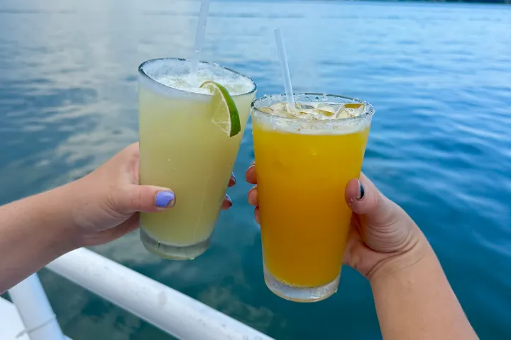 Two hands hold drinks with straws on a boat, overlooking the water and blue sky.