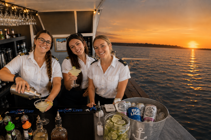 Three women in uniform preparing drinks on a boat at sunset.