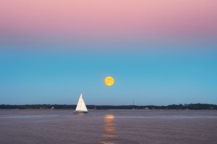 Sailboat on water under a yellow full moon with a pink and blue sky.