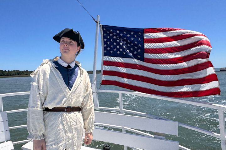 Person in historic attire on boat with American flag waving in background.