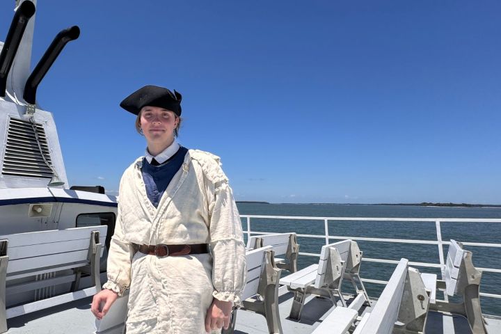 Person in historic attire on a boat deck with clear sky and water in background.