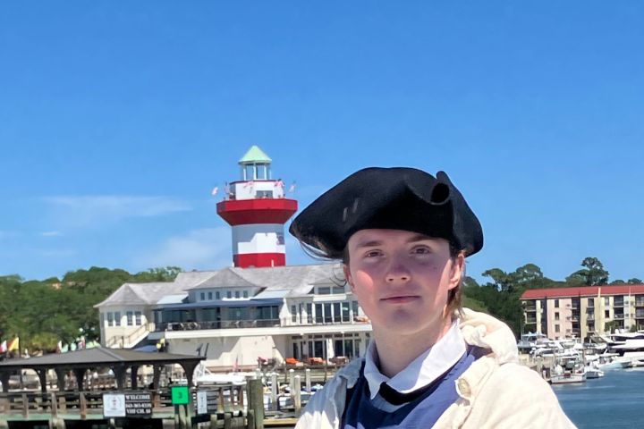 Person in historical attire near a harbor and lighthouse.