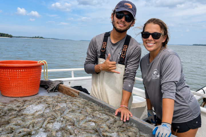 Two people on a boat with a table full of shrimp, smiling at the camera.