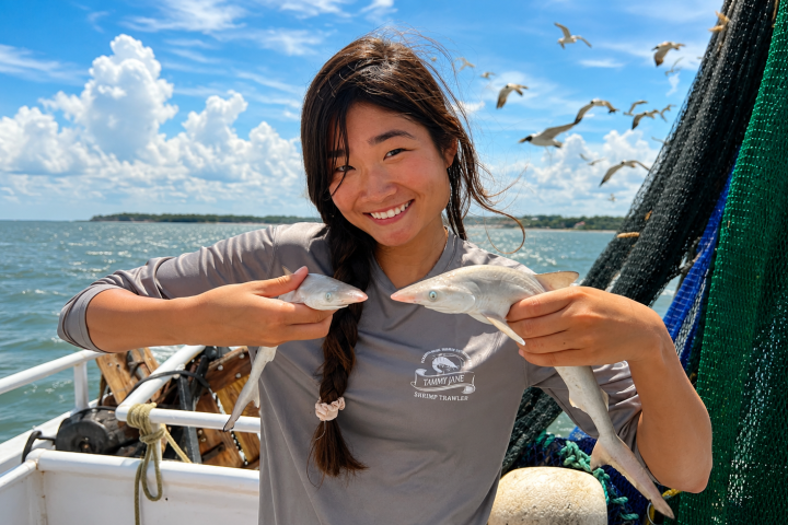 Woman on boat holding fish, smiling, while seagulls fly overhead.