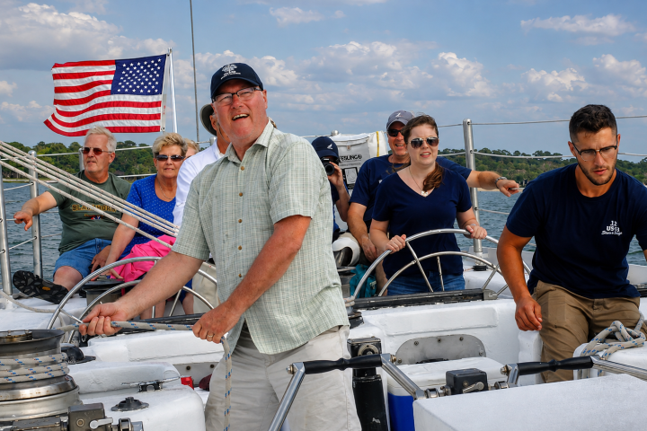 Group sailing on a boat with an American flag, people are smiling and steering.