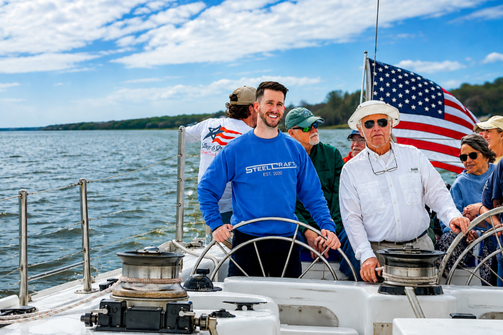 People sailing on a boat with American flag, one man steering, others enjoying the view.